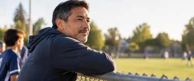 A man watches a youth soccer game from behind a chain-link fence.